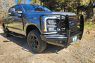 Fab Fours FS23-Q5960-1 Black Steel Elite Full Guard Front Bumper installed on a 2023 Ford F-250 Super Duty, featuring a matte black powder-coated finish, full grille guard, sensor compatibility, and mounts for 3x3 LED cube lights.