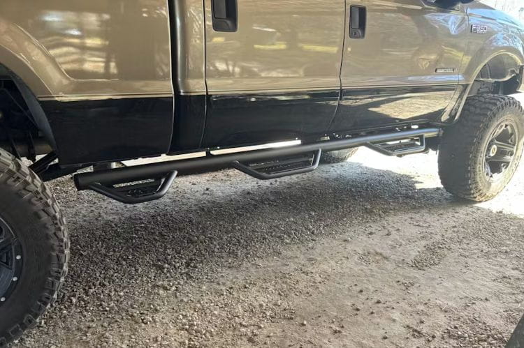 Side view of a vehicle with N-Fab HPF99105CC-6-TX Podium LG stainless steel steps bar for 1999–2016 Ford F250 and F350 Super Duty Crew Cab with single rear wheel and 6'9" bed, durable side step with textured step pads on a gravel surface