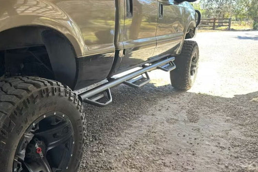 Close-up of a truck with a N-Fab HPF99105CC-6-TX Podium LG stainless steel steps bar for 1999–2016 Ford F250 and F350 Super Duty Crew Cab with single rear wheel and 6'9" bed, durable side step with textured step pads on a gravel surface